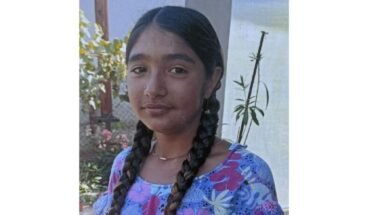 Young girl with two long braids wearing a blue floral dress, standing outdoors near a garden fence.