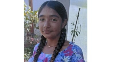 Young girl with two long braids wearing a blue floral dress, standing outdoors near a garden fence.