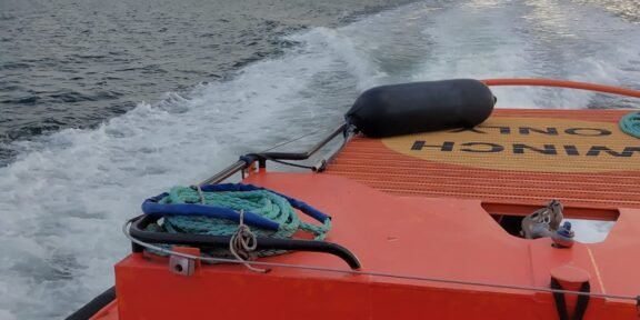 Orange rescue boat with coiled rope on deck, speeding across choppy sea toward the horizon under cloudy sky