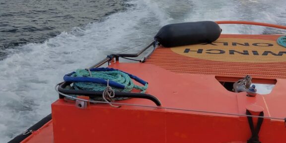 Orange rescue boat with coiled rope on deck, speeding across choppy sea toward the horizon under cloudy sky