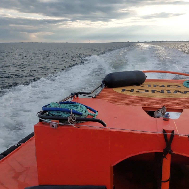 Orange rescue boat with coiled rope on deck, speeding across choppy sea toward the horizon under cloudy sky