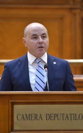 Bald man in a navy suit speaks at a wooden podium in a legislative chamber labeled Camera Deputatilor.