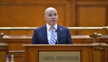 Bald man in a navy suit speaks at a wooden podium in a legislative chamber labeled Camera Deputatilor.