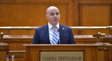 Bald man in a navy suit speaks at a wooden podium in a legislative chamber labeled Camera Deputatilor.