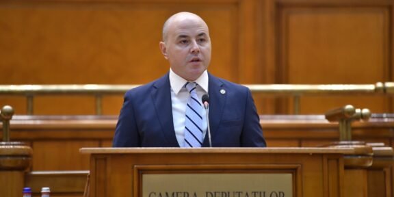Bald man in a navy suit speaks at a wooden podium in a legislative chamber labeled Camera Deputatilor.