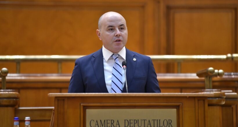 Bald man in a navy suit speaks at a wooden podium in a legislative chamber labeled Camera Deputatilor.