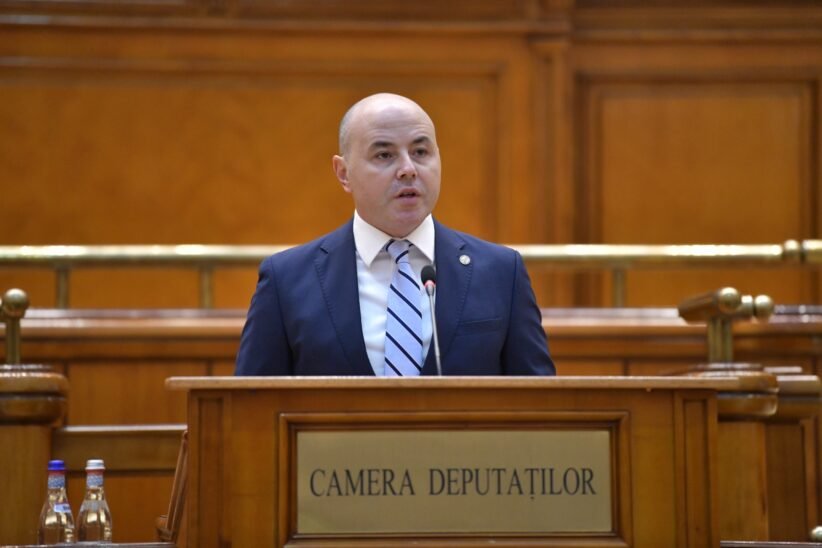 Bald man in a navy suit speaks at a wooden podium in a legislative chamber labeled Camera Deputatilor.
