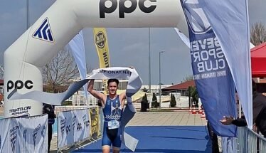 Triathlete in blue suit crosses finish line under an inflatable arch with PPC branding, holding the tape above head, bib number 36 visible on a blue carpeted finish area.