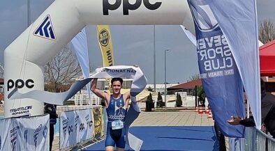 Triathlete in blue suit crosses finish line under an inflatable arch with PPC branding, holding the tape above head, bib number 36 visible on a blue carpeted finish area.
