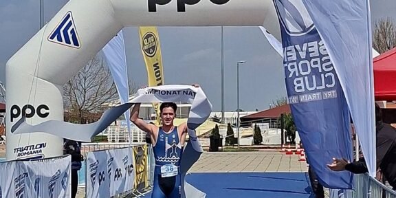 Triathlete in blue suit crosses finish line under an inflatable arch with PPC branding, holding the tape above head, bib number 36 visible on a blue carpeted finish area.