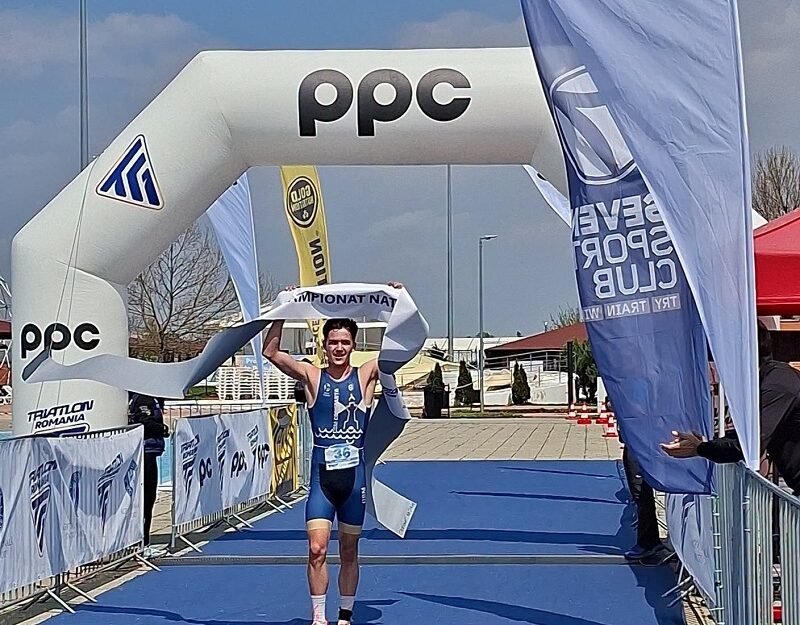 Triathlete in blue suit crosses finish line under an inflatable arch with PPC branding, holding the tape above head, bib number 36 visible on a blue carpeted finish area.