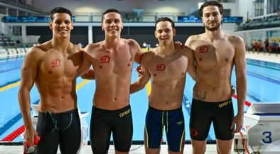 Four male swimmers posing together on an indoor pool deck, wearing black or dark swim briefs with red team logos on their chests.