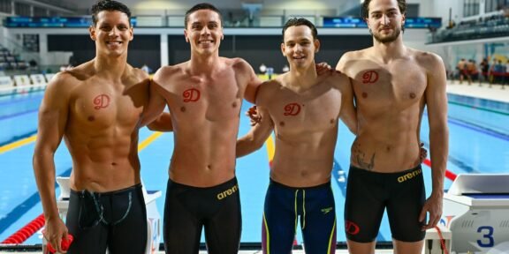 Four male swimmers posing together on an indoor pool deck, wearing black or dark swim briefs with red team logos on their chests.