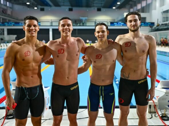 Four male swimmers posing together on an indoor pool deck, wearing black or dark swim briefs with red team logos on their chests.