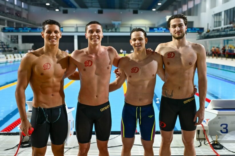Four male swimmers posing together on an indoor pool deck, wearing black or dark swim briefs with red team logos on their chests.