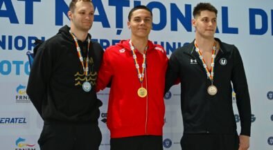 Three male athletes stand on a medals podium, wearing their neck medals and smiling, with a sponsor backdrop behind them.