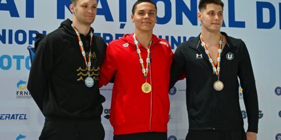 Three male athletes stand on a medals podium, wearing their neck medals and smiling, with a sponsor backdrop behind them.