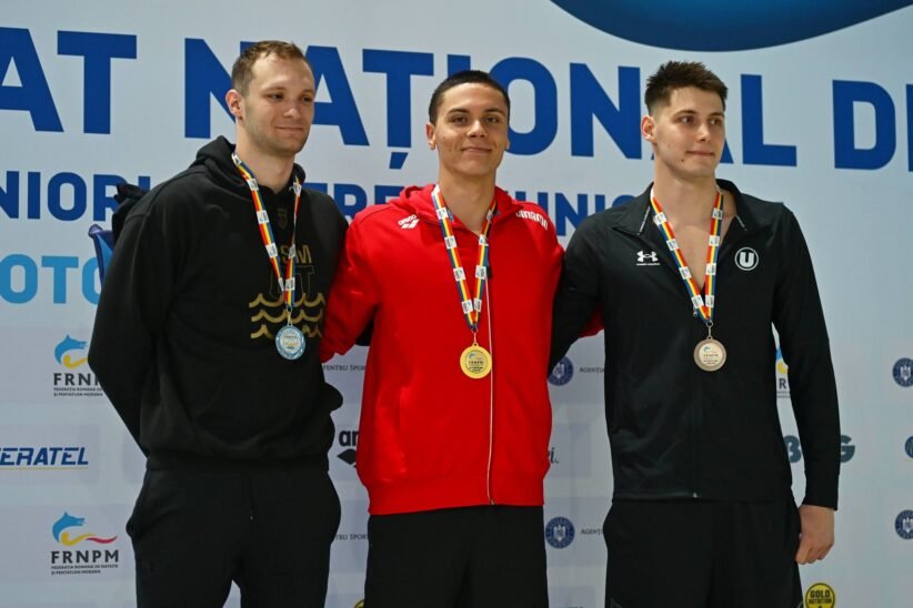 Three male athletes stand on a medals podium, wearing their neck medals and smiling, with a sponsor backdrop behind them.