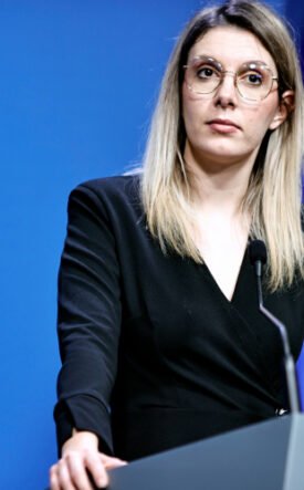 Woman in a black blazer speaks at a podium during a press conference, with a blue backdrop and European flag in the background.