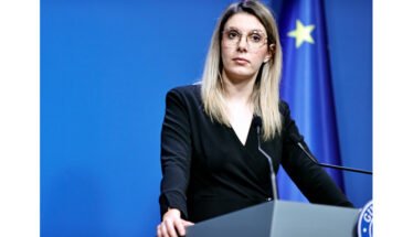 Woman in a black blazer speaks at a podium during a press conference, with a blue backdrop and European flag in the background.