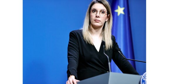 Woman in a black blazer speaks at a podium during a press conference, with a blue backdrop and European flag in the background.