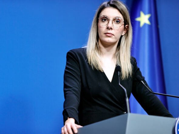 Woman in a black blazer speaks at a podium during a press conference, with a blue backdrop and European flag in the background.