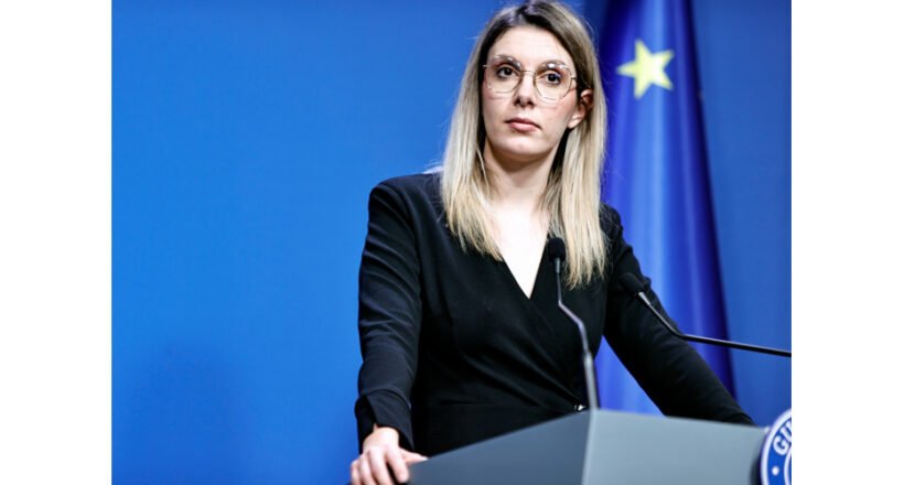 Woman in a black blazer speaks at a podium during a press conference, with a blue backdrop and European flag in the background.
