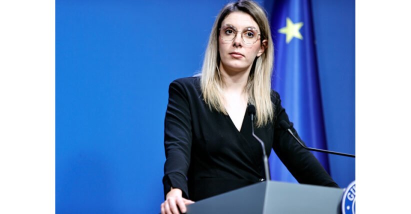 Woman in a black blazer speaks at a podium during a press conference, with a blue backdrop and European flag in the background.