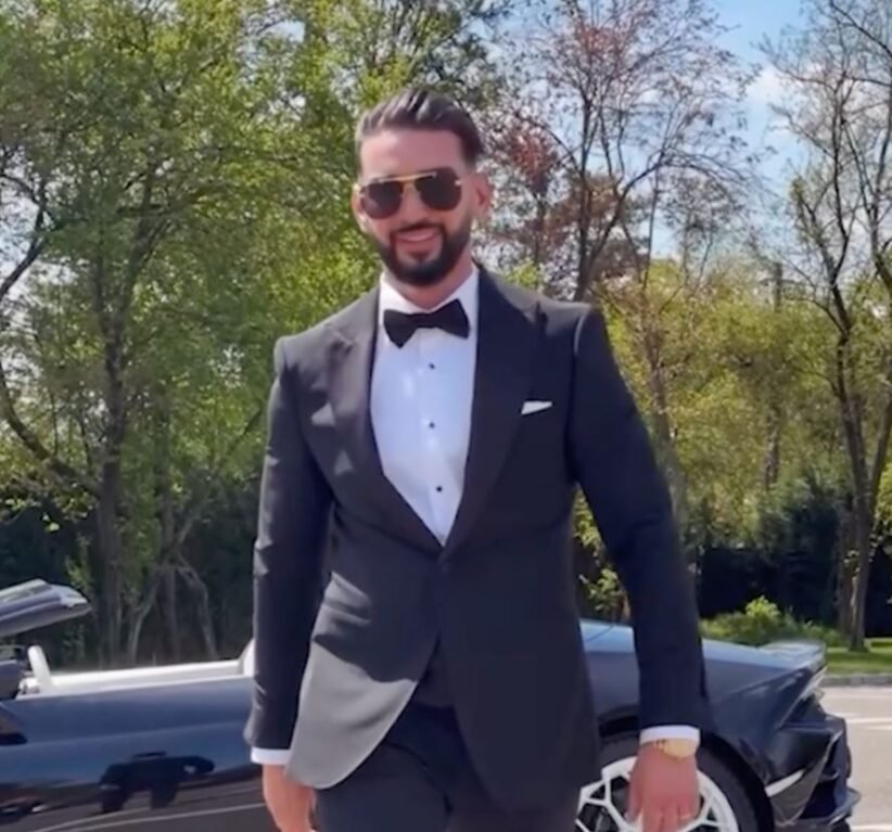 Man in a black tuxedo, white dress shirt, and bow tie standing outdoors with a sports car behind him, wearing sunglasses.