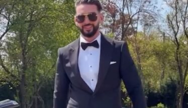 Man in a black tuxedo, white dress shirt, and bow tie standing outdoors with a sports car behind him, wearing sunglasses.