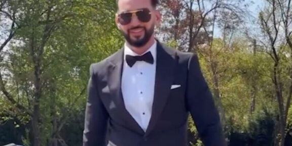 Man in a black tuxedo, white dress shirt, and bow tie standing outdoors with a sports car behind him, wearing sunglasses.
