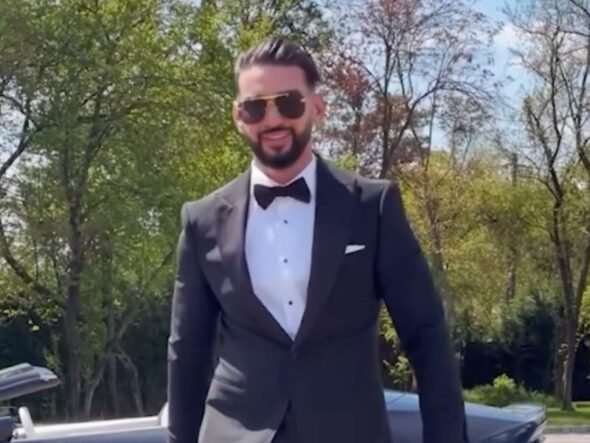 Man in a black tuxedo, white dress shirt, and bow tie standing outdoors with a sports car behind him, wearing sunglasses.