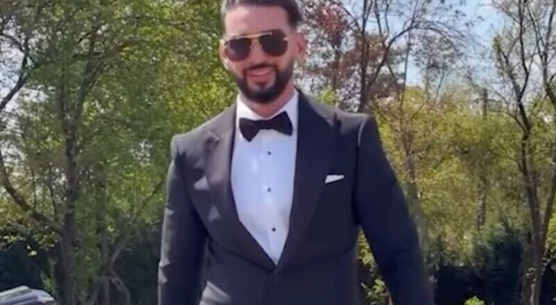 Man in a black tuxedo, white dress shirt, and bow tie standing outdoors with a sports car behind him, wearing sunglasses.
