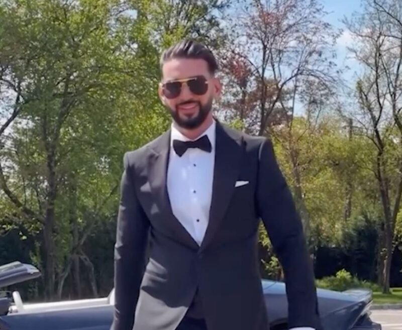 Man in a black tuxedo, white dress shirt, and bow tie standing outdoors with a sports car behind him, wearing sunglasses.