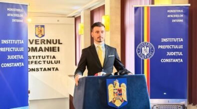Man in a dark suit and yellow tie speaks at a podium with the Romanian coat of arms, flanked by official banners.