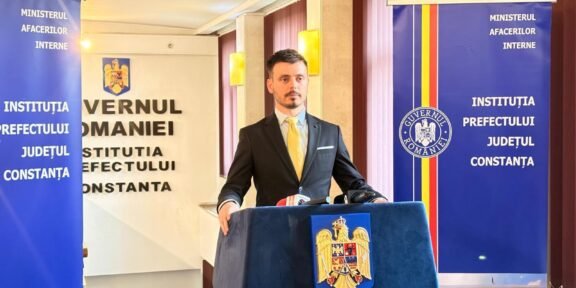 Man in a dark suit and yellow tie speaks at a podium with the Romanian coat of arms, flanked by official banners.