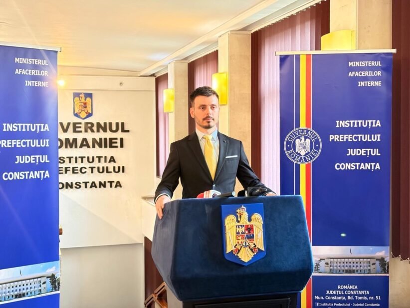 Man in a dark suit and yellow tie speaks at a podium with the Romanian coat of arms, flanked by official banners.