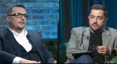 Split-screen interview with two men seated on green couches, one in a dark suit on the left and the other in a gray blazer on the right, talking.