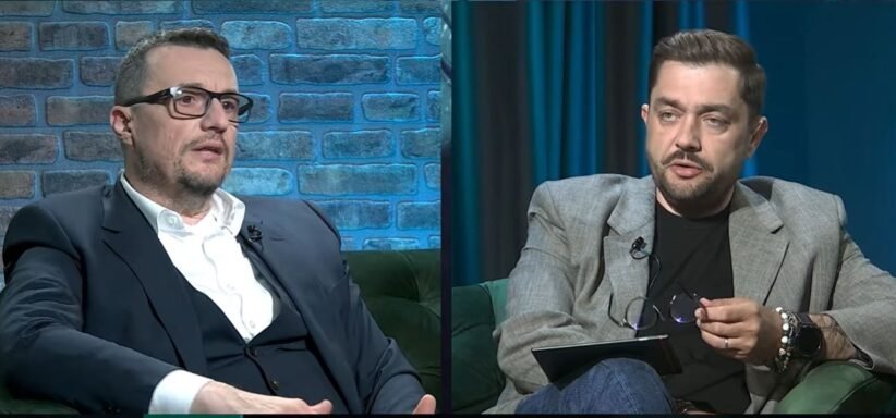 Split-screen interview with two men seated on green couches, one in a dark suit on the left and the other in a gray blazer on the right, talking.