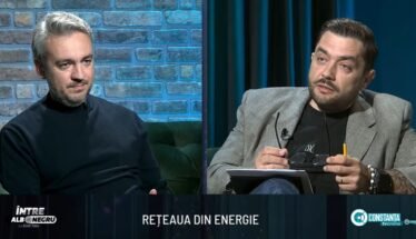 Two men sit on a studio set in conversation; brick wall background on the left and dark curtains on the right.