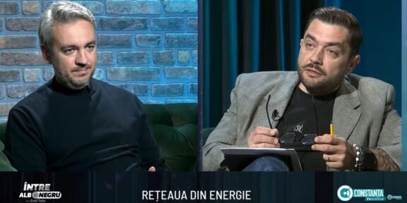 Two men sit on a studio set in conversation; brick wall background on the left and dark curtains on the right.