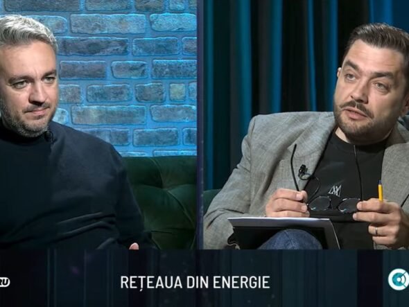 Two men sit on a studio set in conversation; brick wall background on the left and dark curtains on the right.