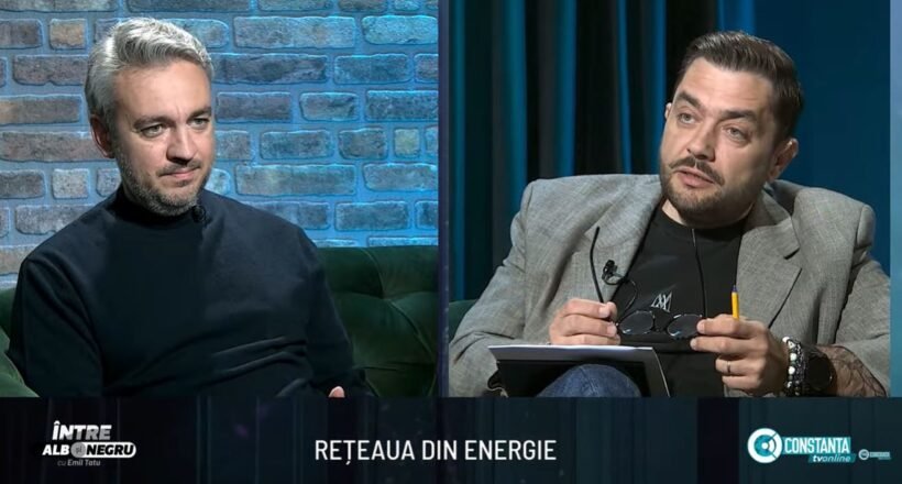 Two men sit on a studio set in conversation; brick wall background on the left and dark curtains on the right.