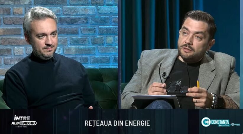 Two men sit on a studio set in conversation; brick wall background on the left and dark curtains on the right.