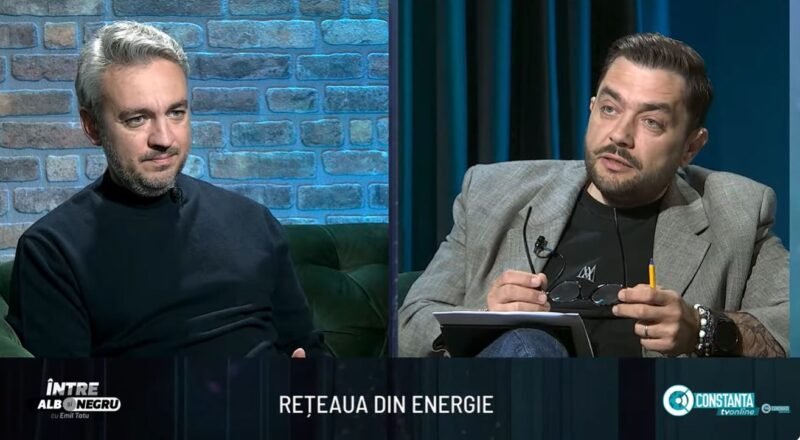 Two men sit on a studio set in conversation; brick wall background on the left and dark curtains on the right.