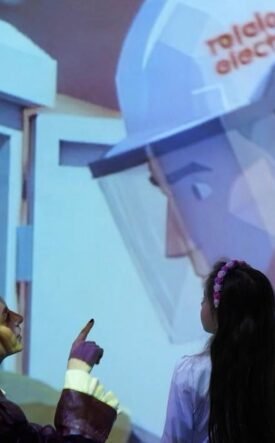 Two people watch and point at a large projection of a healthcare worker in PPE in an exhibit setting.