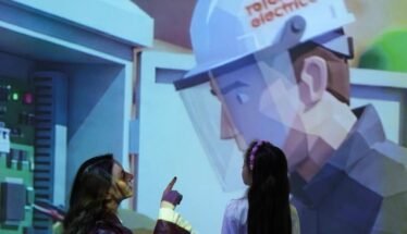 Two people watch and point at a large projection of a healthcare worker in PPE in an exhibit setting.