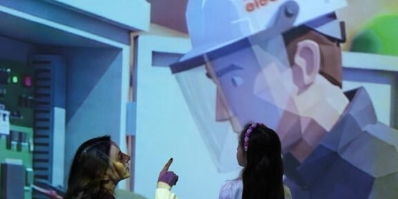 Two people watch and point at a large projection of a healthcare worker in PPE in an exhibit setting.