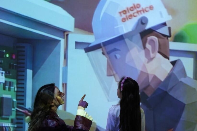 Two people watch and point at a large projection of a healthcare worker in PPE in an exhibit setting.
