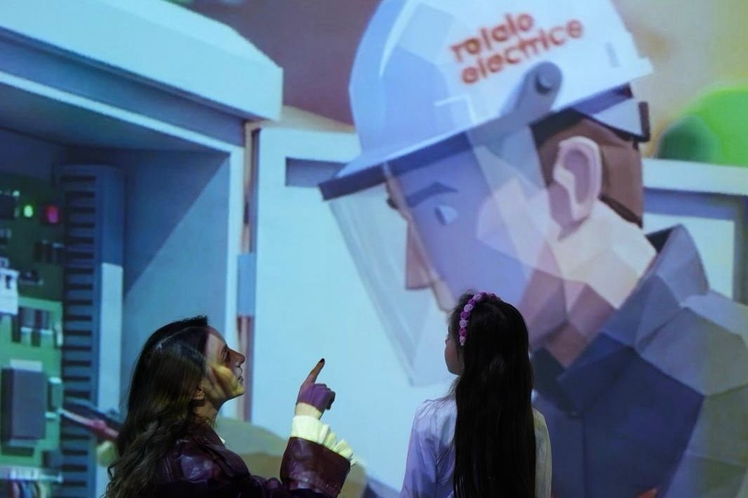 Two people watch and point at a large projection of a healthcare worker in PPE in an exhibit setting.
